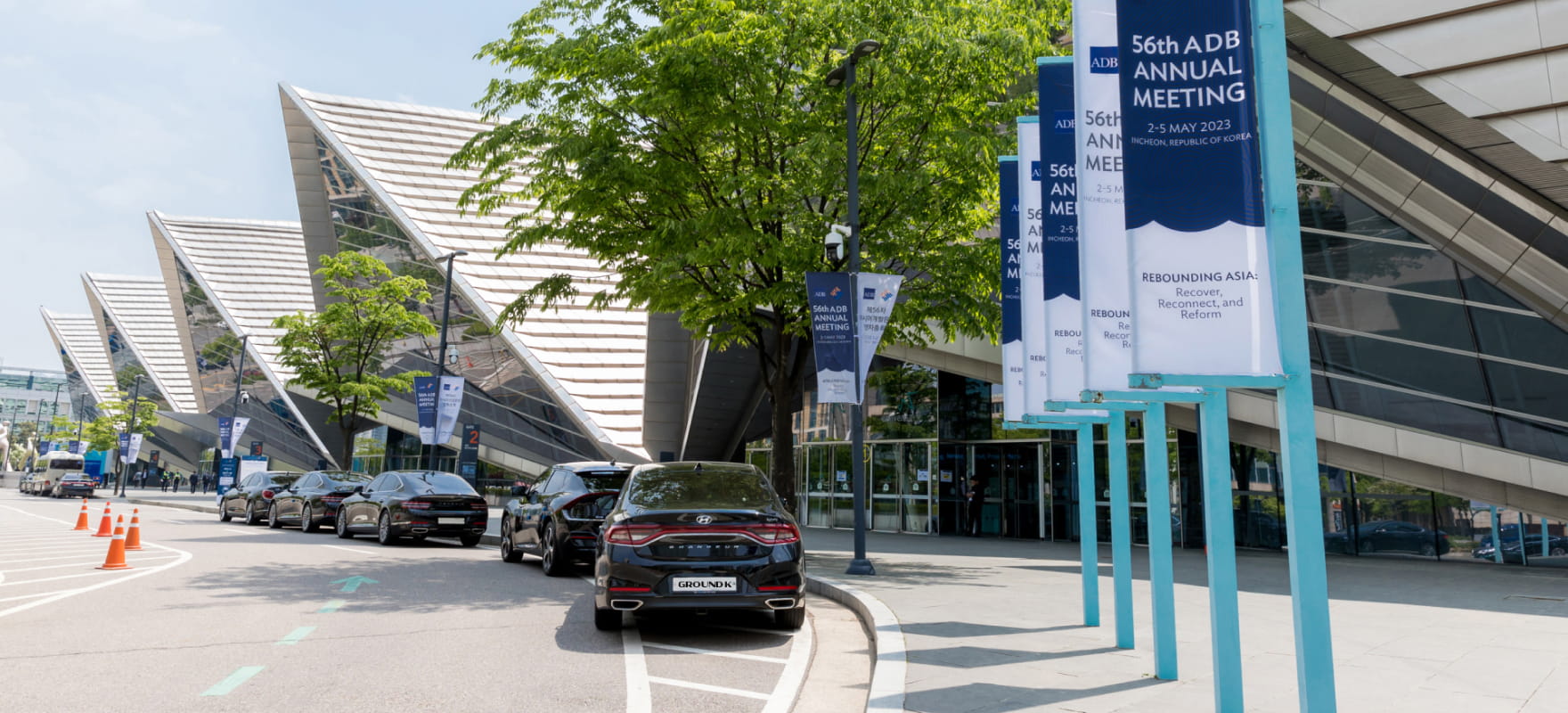 Black protocol vehicles lined up waiting with banners showing '56th Asian Development Bank Annual Meeting' overhead.