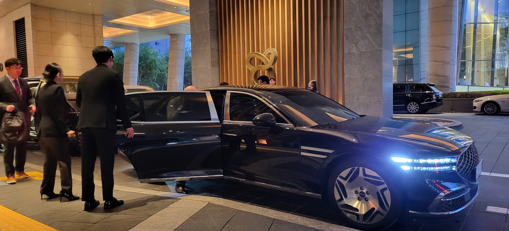 A professional chauffeur in a suit opening the door of a black Genesis G90 luxury sedan for a VIP guest at the AFC awards.