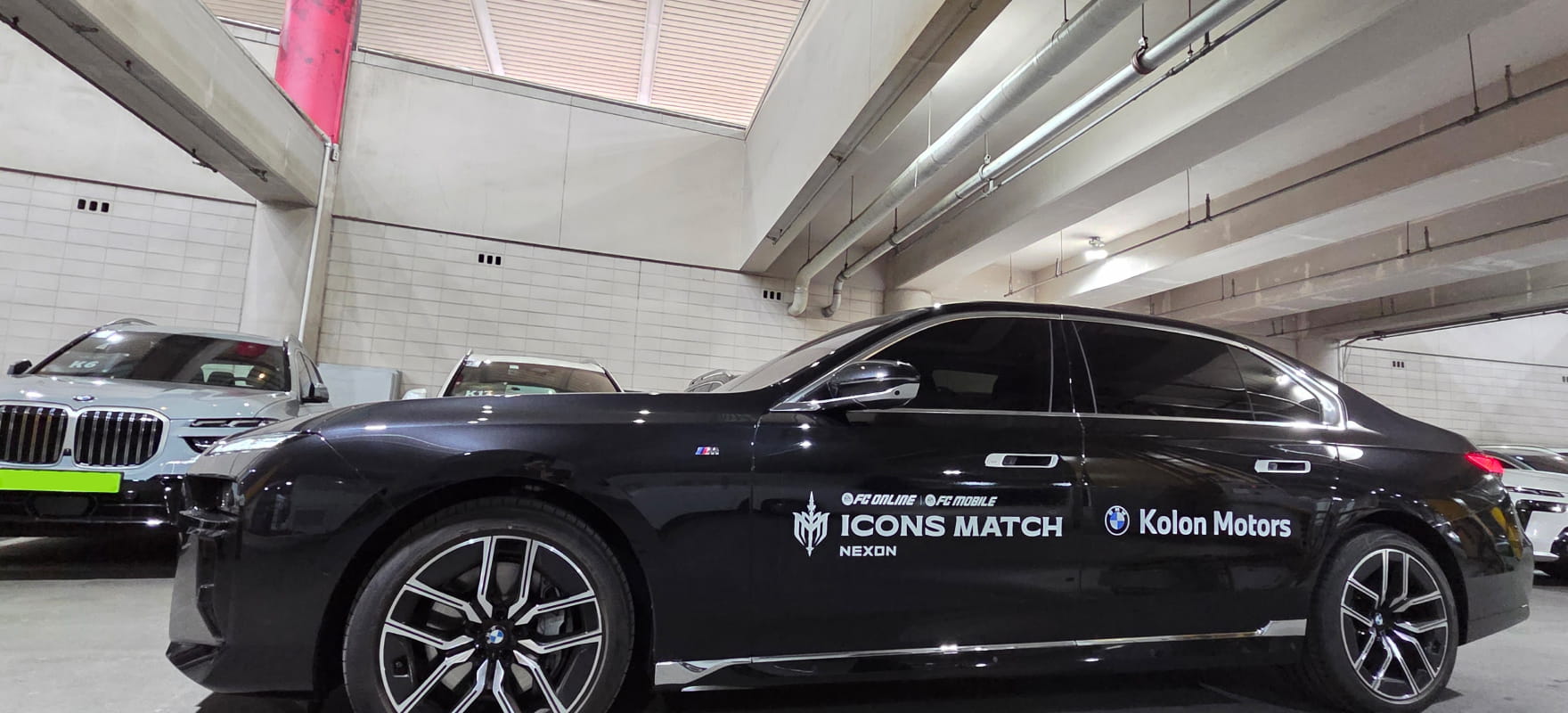 Side profile of a black BMW luxury sedan featuring FC Online ICONS Match Nexon and Kolon Motors decals in a staging area.