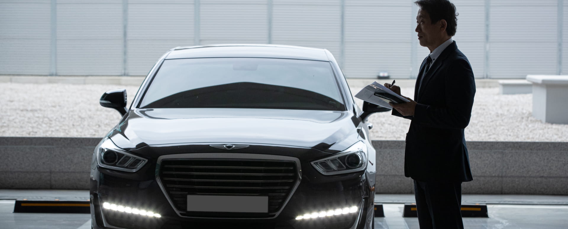 Chauffeur standing in front of a black luxury sedan, holding documents.