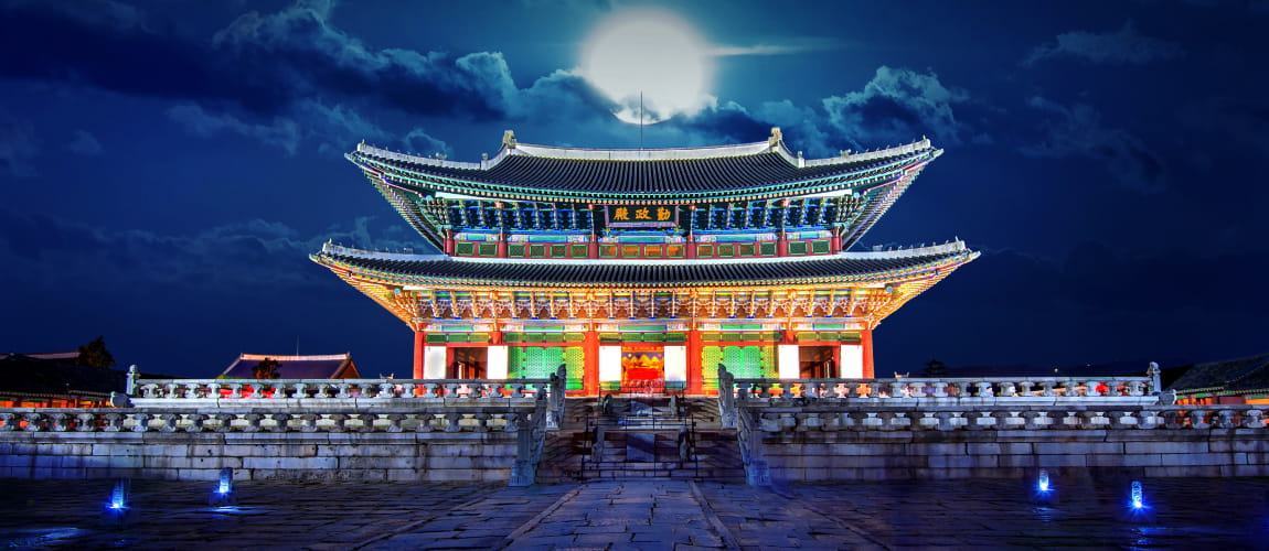 Illuminated Geunjeongjeon Hall of Gyeongbokgung Palace in Seoul, South Korea, beautifully lit at night under a full moon with a dramatic cloudy sky.